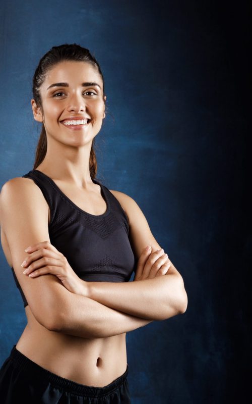 Young beautiful sportive girl posing with crossed arms over dark background. Copy space.